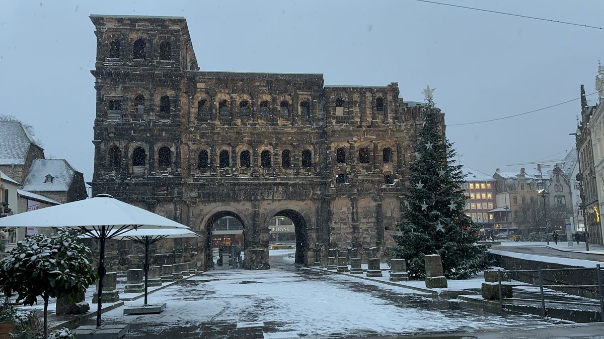 Die Porta Nigra in Trier bei Schneefall im Winter, mit beleuchtetem Weihnachtsbaum auf dem Porta-Nigra-Platz und den Fundamenten der römischen Stadtmauer im Vordergrund